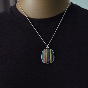 May include: A silver necklace with a square pendant featuring a striped pattern in shades of blue, green, brown, and yellow. The pendant is framed in silver and hangs from a delicate silver chain. The necklace is displayed on a black background.