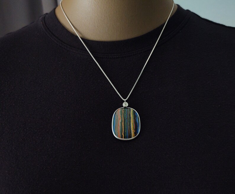 May include: A silver necklace with a square pendant featuring a striped pattern in shades of blue, green, brown, and yellow. The pendant is framed in silver and hangs from a delicate silver chain. The necklace is displayed on a black background.