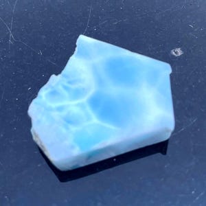 May include: A polished, irregular pentagonal piece of light blue Larimar gemstone. The stone features a distinct marbled pattern with swirling white and sky-blue veins throughout its surface, showcasing a smooth, semi-translucent texture.