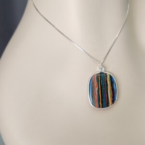May include: A silver necklace featuring a rectangular pendant with a colorful, striped pattern. The pendant has a silver border and is suspended from a delicate silver chain. The striped pattern includes shades of blue, green, red, and yellow.