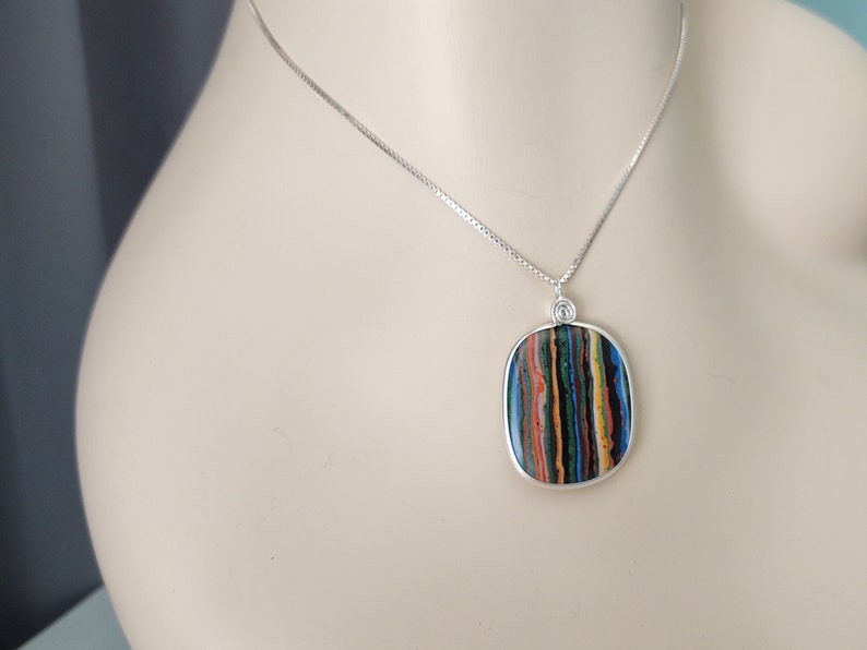 May include: A silver necklace featuring a rectangular pendant with a colorful, striped pattern. The pendant has a silver border and is suspended from a delicate silver chain. The striped pattern includes shades of blue, green, red, and yellow.