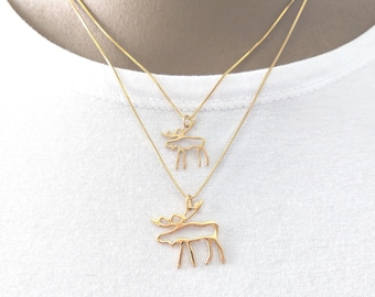May include: Two gold-colored necklaces with moose pendant charms. The moose pendants are outlined in a minimalist style. The necklaces are layered, with one pendant slightly smaller than the other. The background is a white t-shirt.