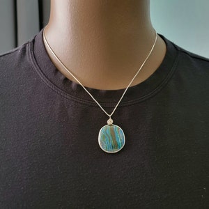 May include: A silver necklace featuring a square pendant with a blue, green, and brown patterned stone. The pendant is approximately 1.5 inches wide and hangs from a delicate silver chain. The necklace is displayed on a black shirt.