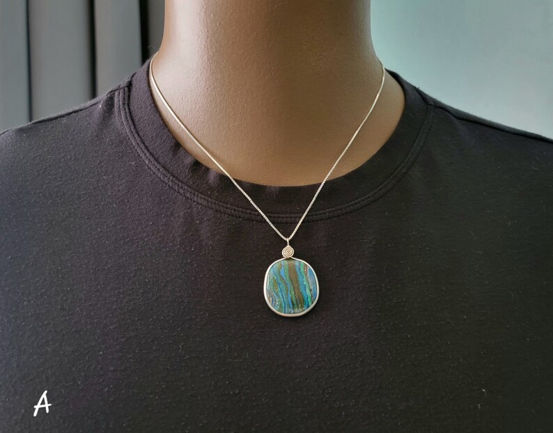 May include: A silver necklace featuring a square pendant with a blue, green, and brown patterned stone. The pendant is approximately 1.5 inches wide and hangs from a delicate silver chain. The necklace is displayed on a black shirt.