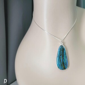 May include: A silver necklace with a pendant featuring a teardrop-shaped, blue and green stone. The stone is encased in a silver frame with a spiral design at the top. The necklace is displayed on a mannequin.