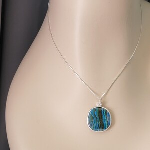 May include: A silver necklace with a square pendant. The pendant features a blue, green, and black marbled design encased in a silver frame. The necklace is displayed on a mannequin.