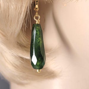 May include: A pair of gold earrings with a dark green faceted gemstone pendant. The earrings are a teardrop shape and have a gold wire wrapped around the top of the gemstone.