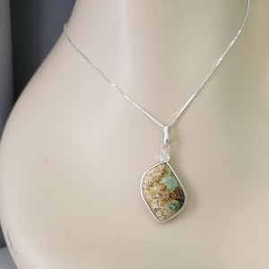 May include: A silver necklace featuring a pendant with an irregular, asymmetrical stone in shades of brown, tan, and teal. The stone is encased in a smooth silver bezel setting with a small spiral detail at the top, suspended from a thin silver chain.