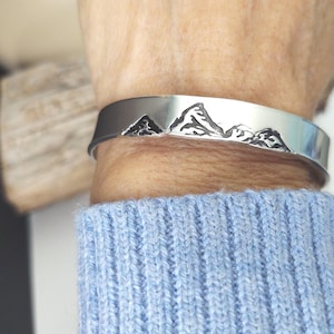 May include: A silver cuff bracelet with a mountain range design. The bracelet is worn on a wrist, and the mountains are etched in black. The person is wearing a light blue sweater.