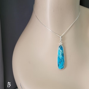 May include: A silver necklace with a long, rectangular pendant. The pendant features a vibrant blue stone with a unique pattern, set in a silver frame. The necklace is displayed on a mannequin.