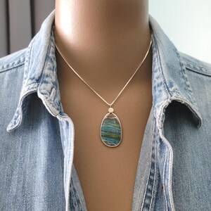 May include: A silver necklace with a pendant featuring a large, oval-shaped stone with blue, green, and brown hues. The pendant is set in a silver frame and hangs from a delicate silver chain. The necklace is worn over a denim shirt.