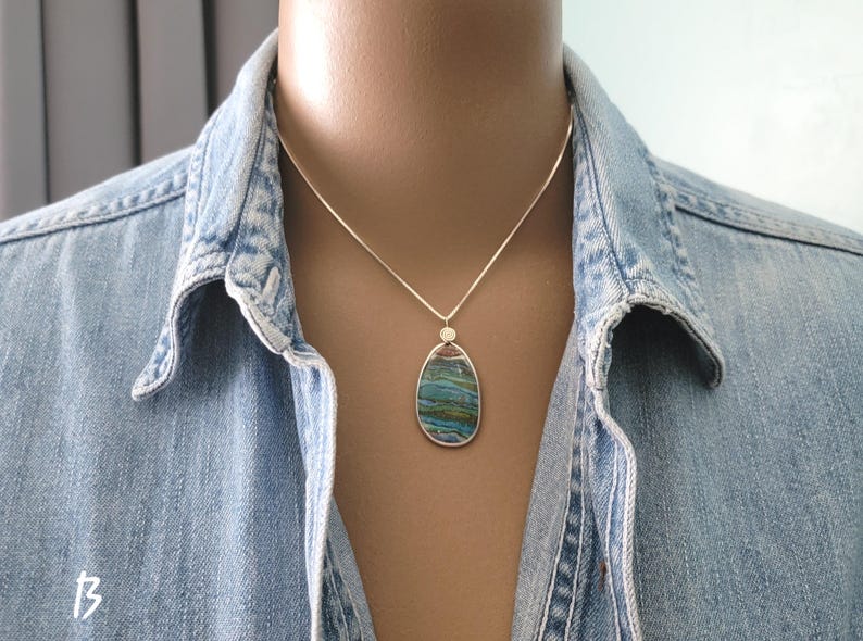 May include: A silver necklace with a pendant featuring a large, oval-shaped stone with blue, green, and brown hues. The pendant is set in a silver frame and hangs from a delicate silver chain. The necklace is worn over a denim shirt.