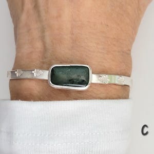May include: A silver cuff bracelet featuring a rectangular, dark green stone set in a smooth bezel. The metal band is decorated with small, stamped fern-like leaf patterns on both sides of the central stone. The letter C is visible.