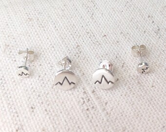 May include: Four silver mountain range stud earrings. Two pairs of earrings are visible, each with a post and a circular design featuring a mountain range graphic. The earrings are displayed on a textured, off-white surface.