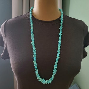 May include: A turquoise blue gemstone necklace made of small, irregular shaped beads. The necklace is long and hangs down to the chest.