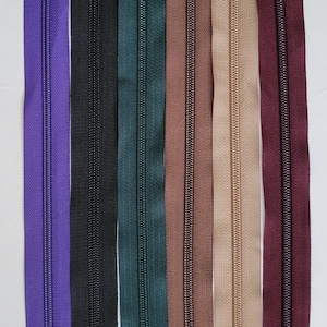 Puede incluir: Una colección de cremalleras verticales en varios colores: morado, negro, verde azulado, marrón, beige y burdeos. Cada cremallera se muestra verticalmente sobre un fondo blanco, mostrando las diferentes opciones de color.
