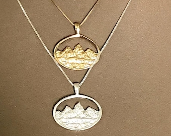 Könnte beinhalten: Zwei Halsketten mit Anhängern im Bergdesign. Ein Anhänger ist goldfarben, der andere silberfarben. Beide Anhänger sind oval und zeigen eine Bergketten-Silhouette. Die Ketten sind filigran und vor braunem Hintergrund.