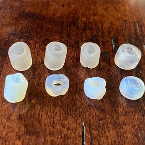 May include: Eight small, translucent, cylindrical and ring-shaped beads in varying shades of white and pale blue. The beads are arranged on a dark brown wooden surface. Some beads have a slightly rough texture.