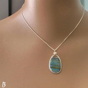 May include: A silver necklace with a pendant. The pendant is an oval-shaped stone with blue, green, and brown hues. The stone is set in a silver bezel and hangs from a delicate silver chain. The necklace is displayed on a person.