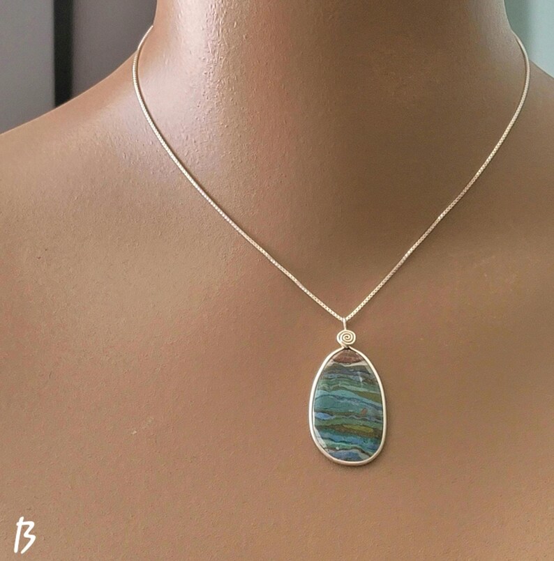May include: A silver necklace with a pendant. The pendant is an oval-shaped stone with blue, green, and brown hues. The stone is set in a silver bezel and hangs from a delicate silver chain. The necklace is displayed on a person.