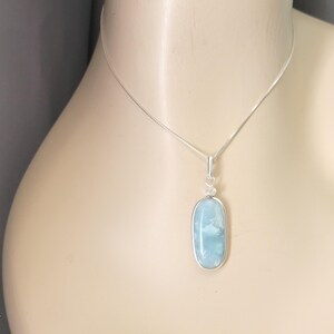 May include: A silver necklace featuring a smooth, oval-shaped light blue stone pendant. The stone is encased in a simple silver wire wrap setting and hangs from a thin, delicate silver chain. The letter A is visible in the bottom left corner.