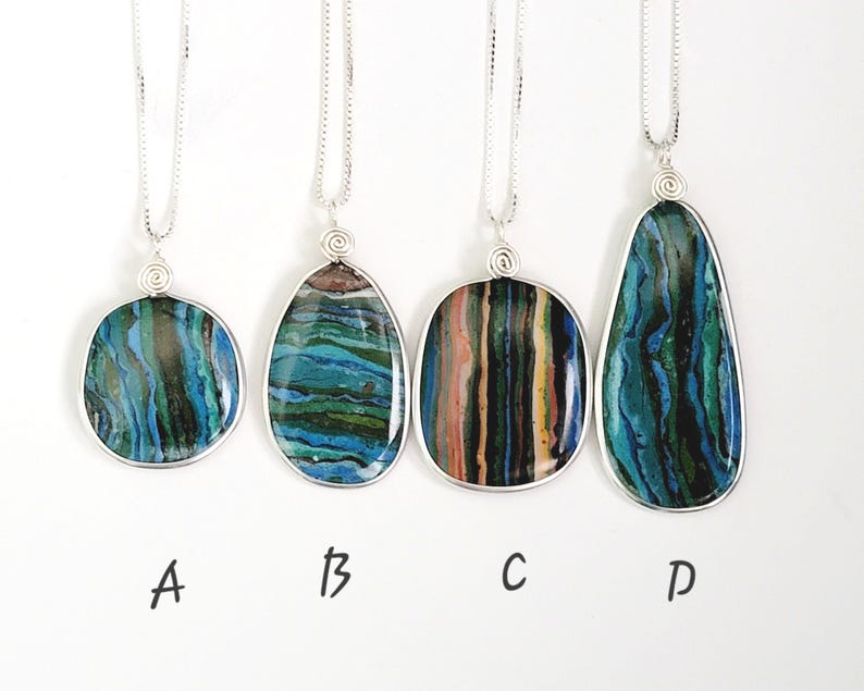 May include: Four silver pendant necklaces with unique, colorful stone pendants. The pendants are various shapes: round, oval, rectangular, and teardrop. Each stone features blue, green, and brown hues. The necklaces are displayed against a white background.