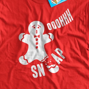 Op de afbeelding: Rood t-shirt met een witte afbeelding van een peperkoeken man die in twee&euml;n breekt, met de witte woorden "OOOOHHH SNAP". Een blauw label met een roze ijslogo is aan het shirt bevestigd.