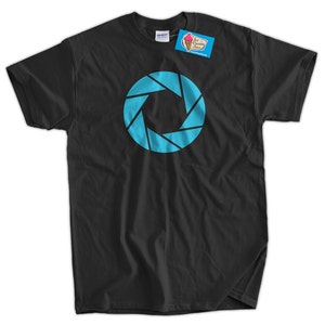 Może przedstawiać: Czarny T-shirt z niebieskim logo Aperture Science. Logo to okrąg z szeregiem niebieskich tr&oacute;jkąt&oacute;w skierowanych do wewnątrz.