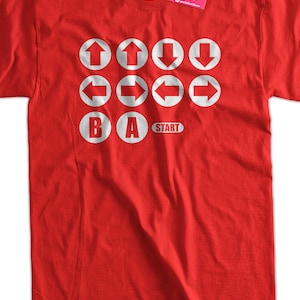 May include: Red t-shirt with a white graphic of a video game controller. The graphic includes the directional buttons, A, B, and Start.