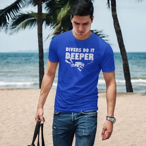 May include: A man wearing a royal blue t-shirt with the text "Divers Do It Deeper" and a white silhouette of a scuba diver. He is wearing blue jeans and a silver watch. He is walking on a sandy beach with a black bag in his hand.
