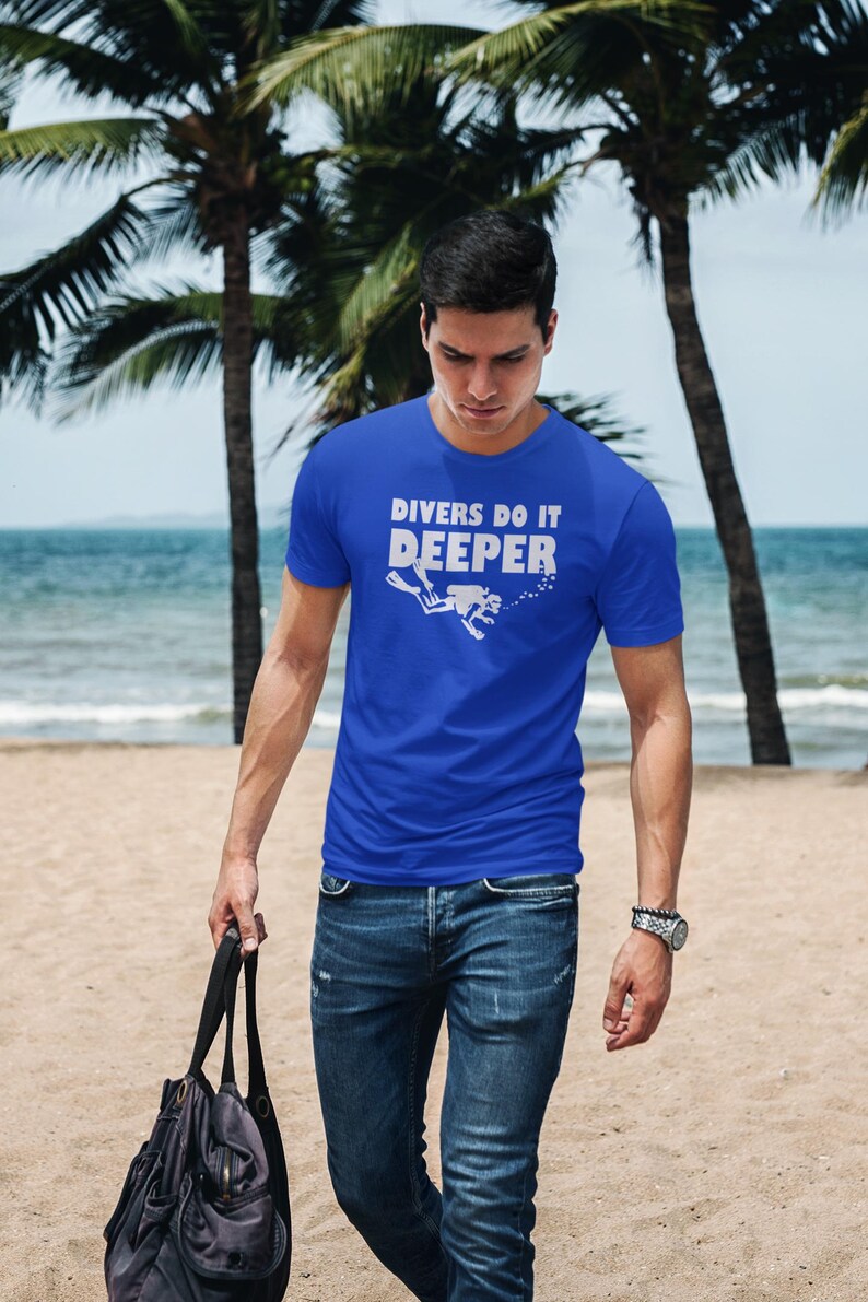 May include: A man wearing a royal blue t-shirt with the text "Divers Do It Deeper" and a white silhouette of a scuba diver. He is wearing blue jeans and a silver watch. He is walking on a sandy beach with a black bag in his hand.