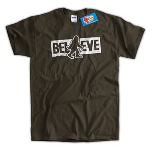 May include: A dark green t-shirt featuring a white graphic of a Bigfoot walking in front of a sign that reads "BELIEVE".