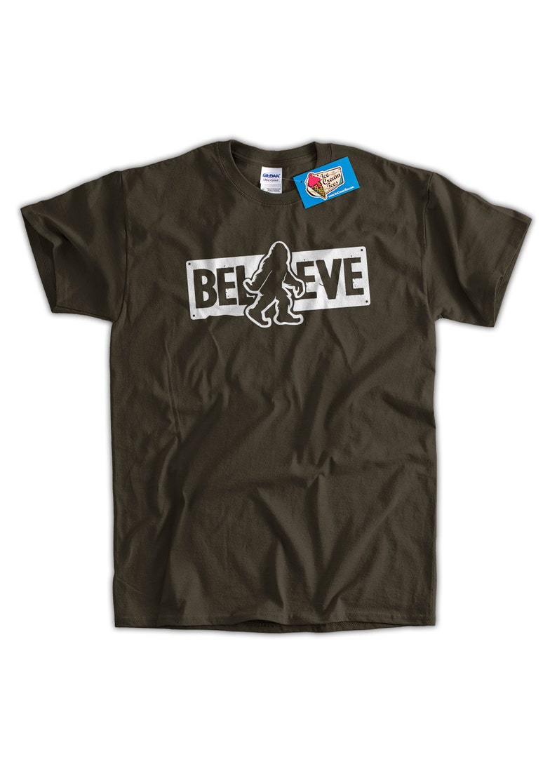 May include: A dark green t-shirt featuring a white graphic of a Bigfoot walking in front of a sign that reads "BELIEVE".