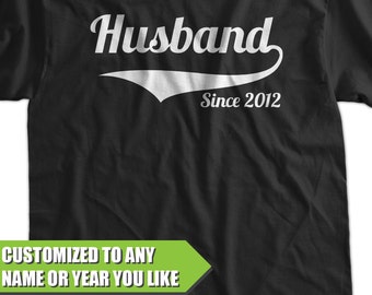 May include: Black t-shirt with white text that reads "Husband Since 2012". The text is in a retro-style font with a white baseball-style arc underneath. The shirt is customizable to any name or year.