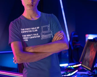 Może przedstawiać: Młoda osoba w niebieskim t-shircie z białym nadrukiem, na którym widnieje napis "The first rule of computer club you don't talk about computer club". Nadruk zawiera również retro komputer.