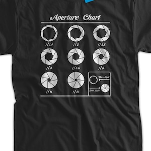 May include: Black t-shirt featuring a white graphic of an aperture chart displaying various f-stops and the corresponding aperture blades. The text "Aperture Chart" is at the top of the graphic. The graphic also includes arrows indicating "More Light" and "Less Light".