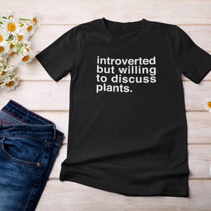 K&ouml;nnte beinhalten: Schwarzes T-Shirt mit wei&szlig;em Text, der "introvertiert, aber bereit, &uuml;ber Pflanzen zu diskutieren" lautet.