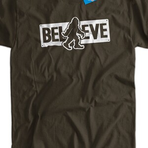 May include: A brown t-shirt with a white graphic of a Bigfoot walking in front of a sign that says "BELIEVE".
