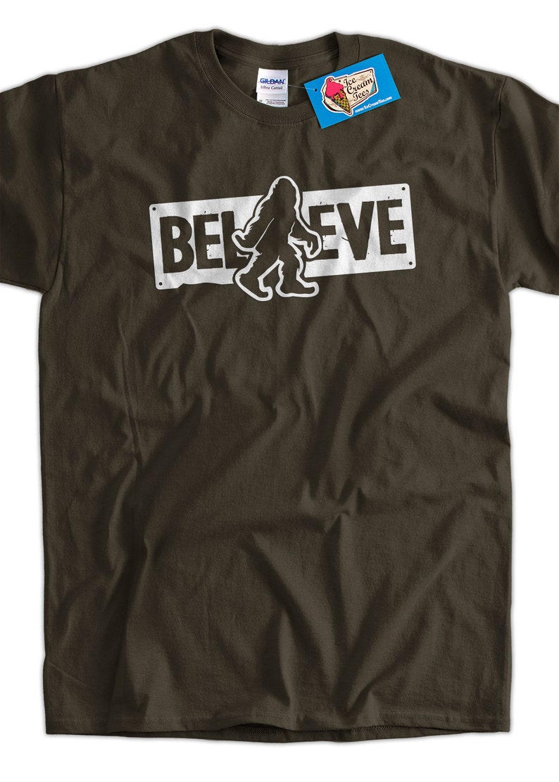 May include: A brown t-shirt with a white graphic of a Bigfoot walking in front of a sign that says "BELIEVE".