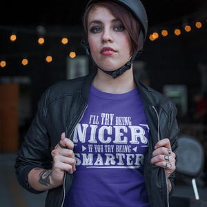 May include: A woman wearing a purple t-shirt with the text "I'll try being nicer if you try being smarter". She is wearing a black leather jacket and a black helmet.