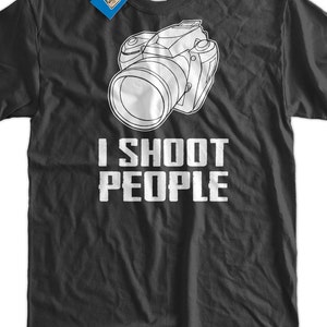May include: Dark gray t-shirt with a white outline of a camera and the text "I SHOOT PEOPLE" in bold, white letters. A blue tag with text hangs from the collar. A fun gift for photographers.