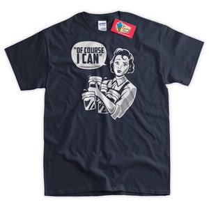 May include: A navy blue t-shirt with a white graphic of a woman holding multiple cups and a speech bubble that says "Of course I can".