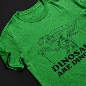 May include: Green t-shirt featuring a black line drawing of two dinosaurs inside a circle. The text "DINOSAURS ARE DINOS" is printed in black below the dinosaurs. The shirt is made of a soft, textured fabric.