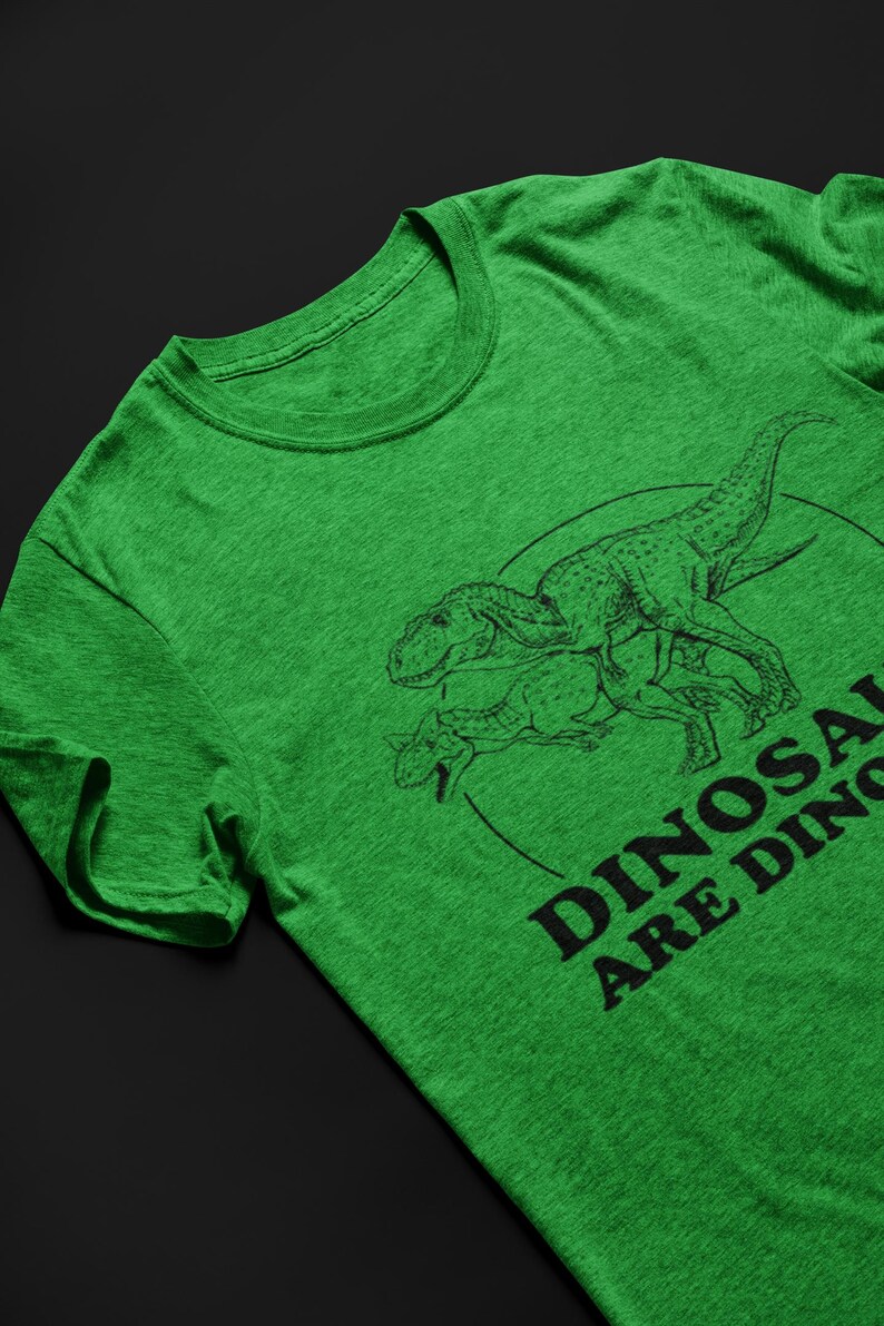 May include: Green t-shirt featuring a black line drawing of two dinosaurs inside a circle. The text "DINOSAURS ARE DINOS" is printed in black below the dinosaurs. The shirt is made of a soft, textured fabric.