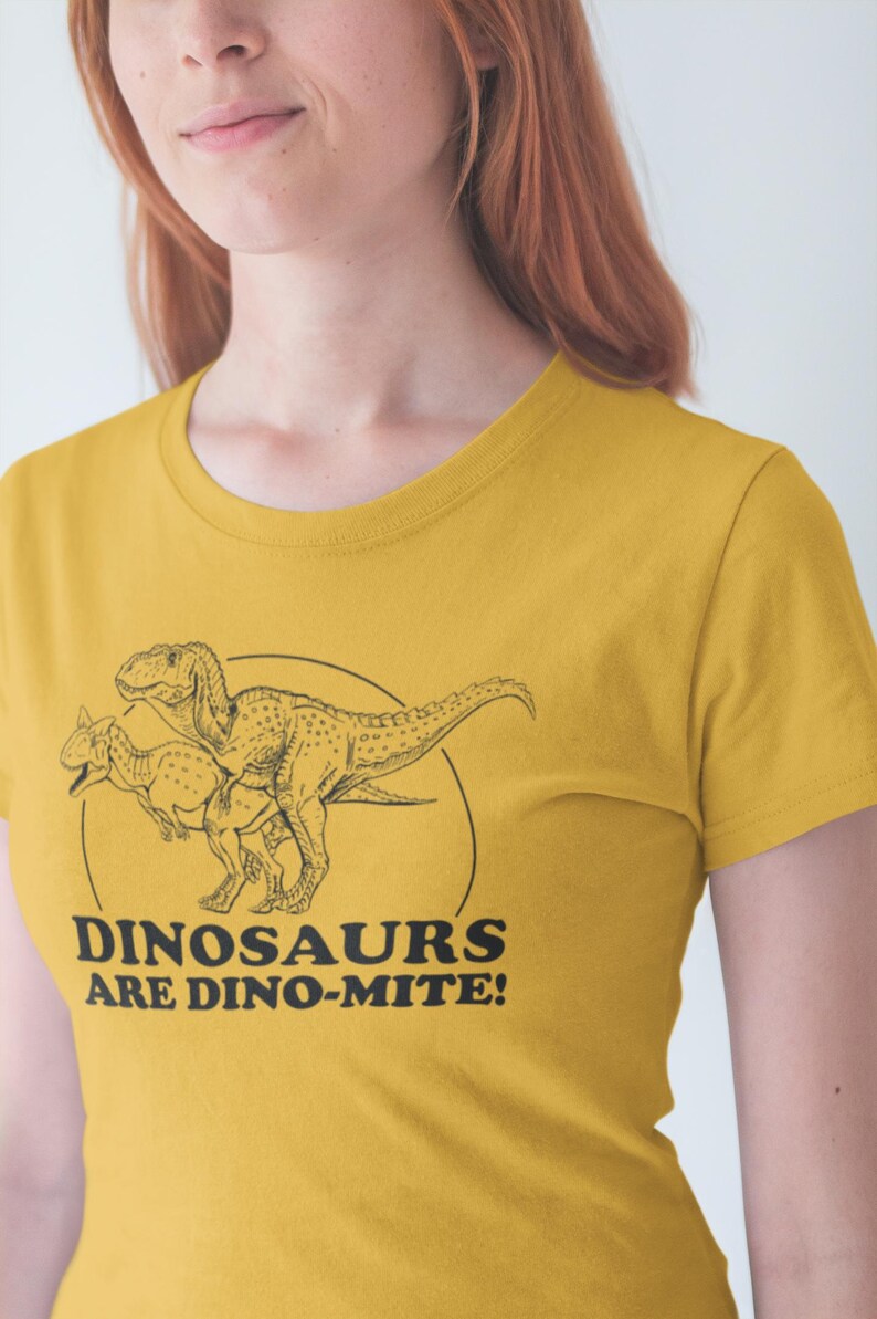 May include: Mustard yellow t-shirt featuring a black line drawing of two dinosaurs inside a circle. The text "DINOSAURS ARE DINO-MITE!" is below the dinosaurs. The shirt is made of a soft material.
