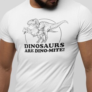 May include: White t-shirt featuring a black and white illustration of two dinosaurs within a circle. The text "DINOSAURS ARE DINO-MITE!" is printed below the illustration. The shirt is made of a soft, comfortable material.