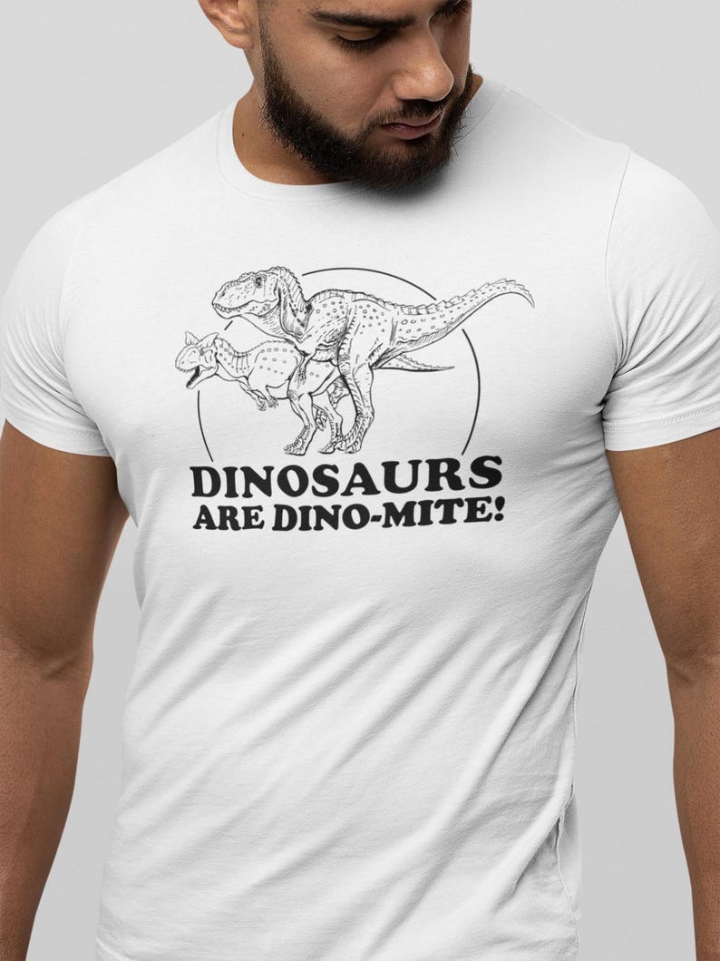May include: White t-shirt featuring a black and white illustration of two dinosaurs within a circle. The text "DINOSAURS ARE DINO-MITE!" is printed below the illustration. The shirt is made of a soft, comfortable material.