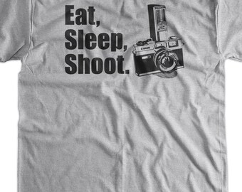 May include: Gray t-shirt with black text that reads "Eat, Sleep, Shoot." A black and white illustration of a camera with a flash is to the right of the text.
