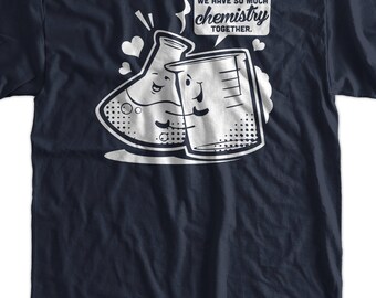May include: A navy blue t-shirt featuring a white graphic of two beakers with faces, one with a flask and the other with a graduated cylinder. The text "We have so much chemistry together." is printed above the beakers.