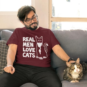 May include: A man wearing a maroon t-shirt with the text "REAL MEN LOVE CATS" and a white cat graphic. He is sitting on a couch and petting a tabby cat.